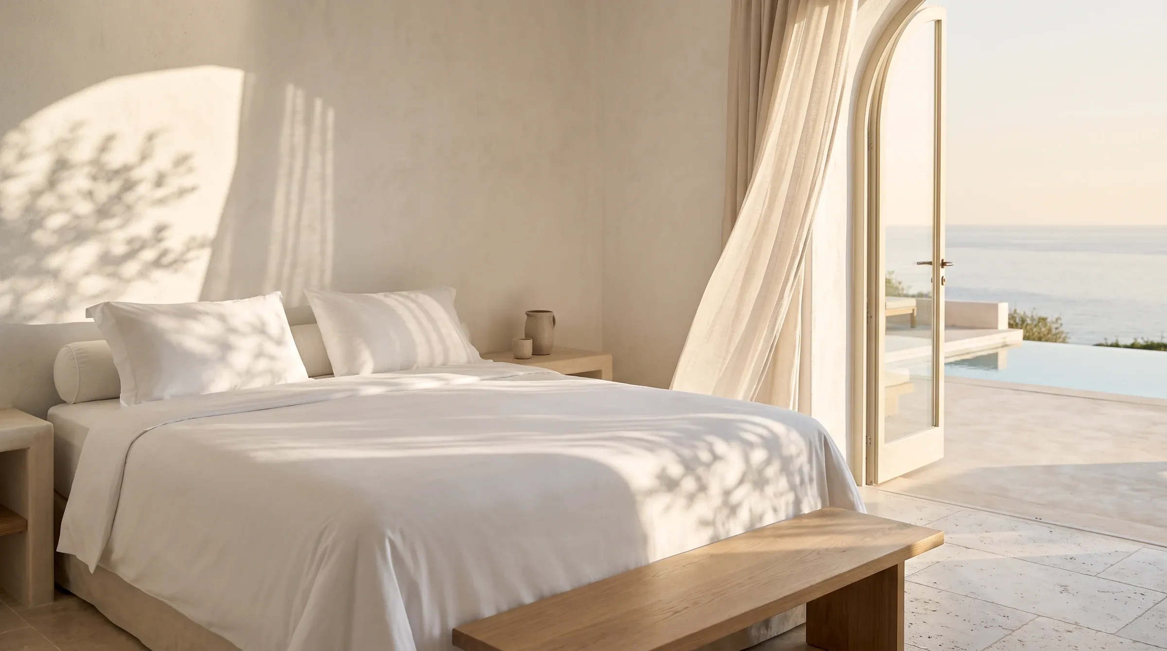 Chambre baignée de lumière du matin, lit blanc fait avec soin, ombres de feuilles d'olivier sur le mur et vue sur la mer